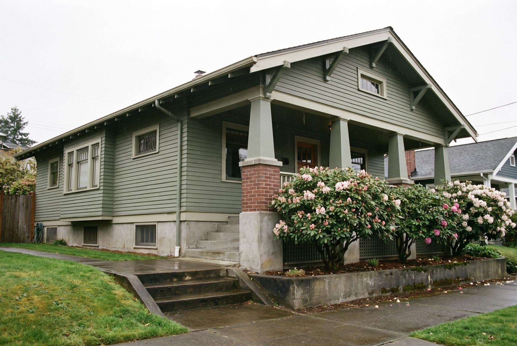 A 1920s craftsman bungalow in Seattle with painted green siding and brick porch piers, its raised concrete foundation and low basement windows visible above a wet sidewalk