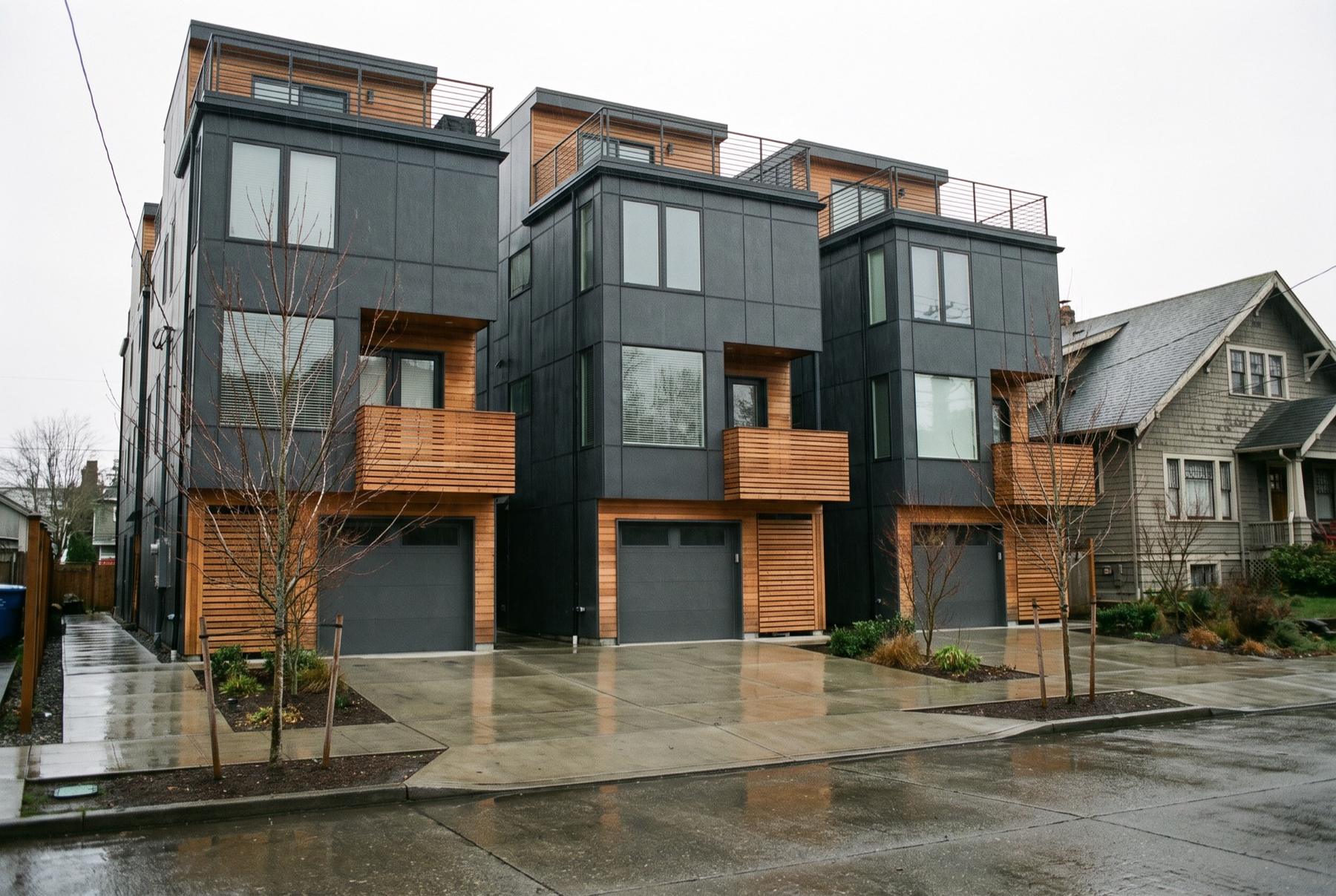 A row of new three-story Seattle townhomes with dark metal siding, cedar balconies and roof decks, garages opening onto a shared concrete driveway, an older craftsman house next door on a rainy afternoon